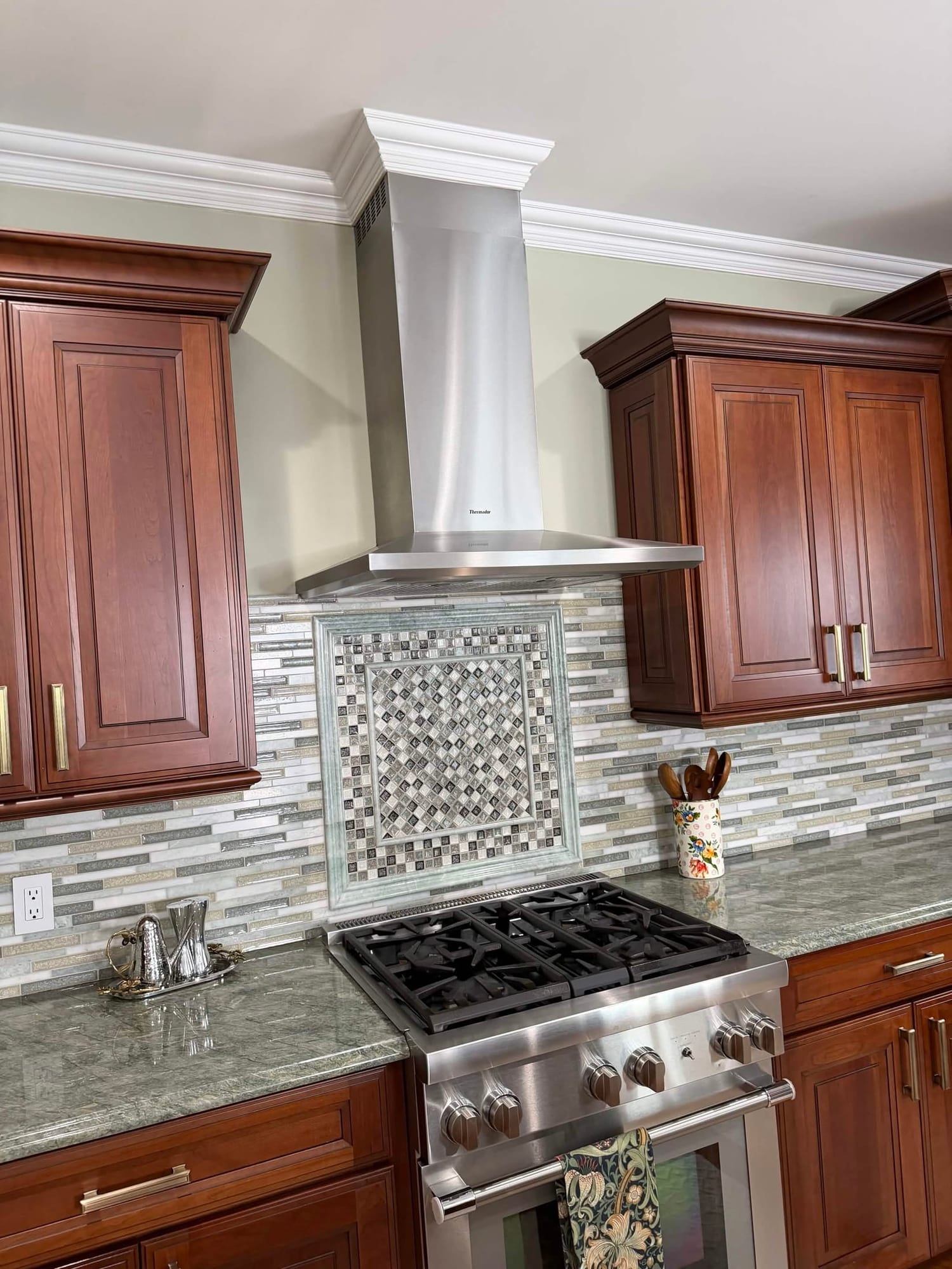 Cherry kitchen with stainless range hood, gas cooktop and a mosaic tile backsplash medallion